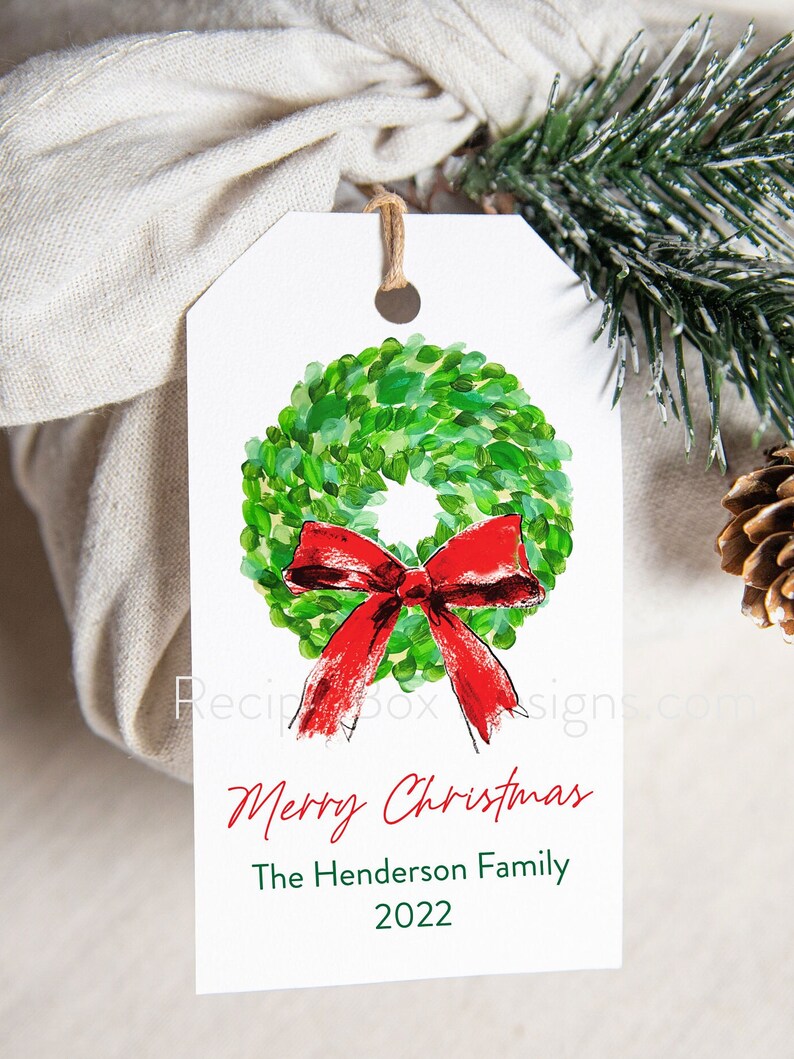 May include: A white gift tag with a watercolor wreath and red bow. The tag reads "Merry Christmas" and "The Henderson Family 2022". A twine string is attached to the tag.
