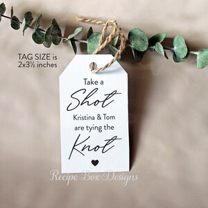 May include: White wedding favor tag with black text that reads "Take a Shot Kristina & Tom are tying the Knot" with a small black heart at the bottom. The tag is 2 by 3 1/2 inches.