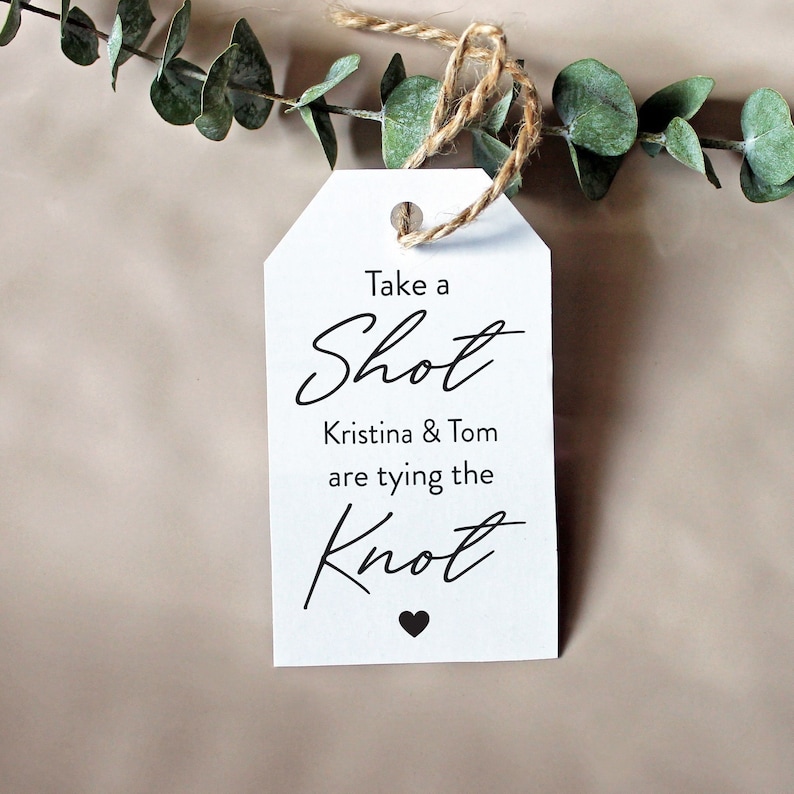 May include: White tag with black text that reads "Take a Shot Kristina & Tom are tying the Knot" with a small black heart at the bottom.