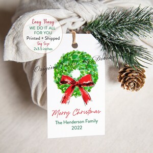 May include: A white gift tag with a watercolor wreath and red bow design. The tag reads "Merry Christmas" and "The Henderson Family 2022". The tag is 2x3.5 inches and is attached to a wrapped gift with twine.