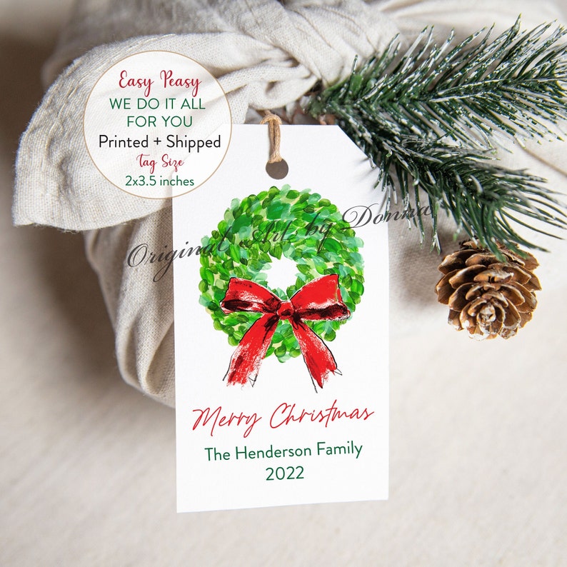 May include: A white gift tag with a watercolor wreath and red bow design. The tag reads "Merry Christmas" and "The Henderson Family 2022". The tag is 2x3.5 inches and is attached to a wrapped gift with twine.