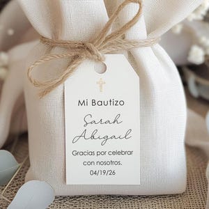 May include: A small, off-white fabric bag tied with twine, featuring a tag with the text "Mi Bautizo Sarah Abigail" and a cross symbol. The tag also includes the text "Gracias por celebrar con nosotros. 04/19/26".