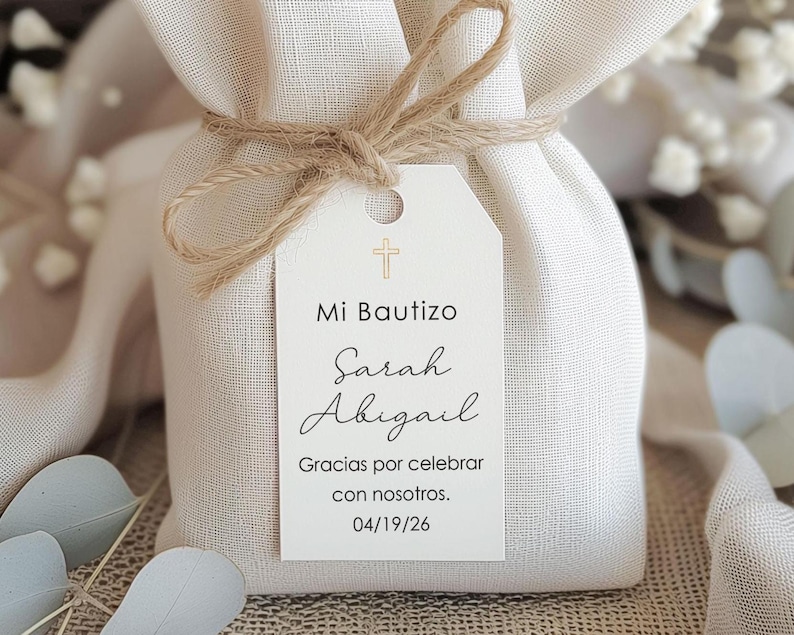 May include: A small, off-white fabric bag tied with twine, featuring a tag with the text "Mi Bautizo Sarah Abigail" and a cross symbol. The tag also includes the text "Gracias por celebrar con nosotros. 04/19/26".