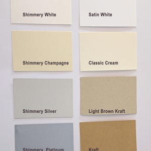May include: A collection of rectangular paper swatches in various colors, including white, cream, silver, and brown. Each swatch is labeled with its color name, such as "Shimmery White" and "Kraft". The arrangement is neat and organized, showcasing the range of paper options.