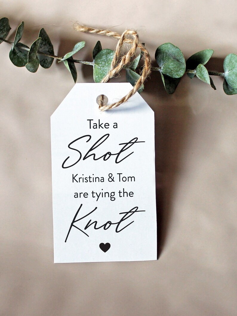 May include: White tag with a brown twine string attached. The tag says "Take a Shot Kristina & Tom are tying the Knot" with a small black heart at the bottom.