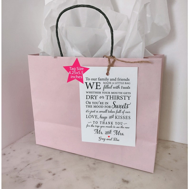 May include: A pink gift bag with a white tag attached. The tag has black text that reads "To our family and friends WE made a little bag filled with treats WHETHER YOUR MOUTH GETS DRY or THIRSTY OR YOU'RE IN THE MOOD FOR Sweets it's just a small token full of our LOVE, hugs and KISSES ~ TO THANK YOU ~ for the trip you made to see the new Mr. and Mrs. Greg and Elsa".