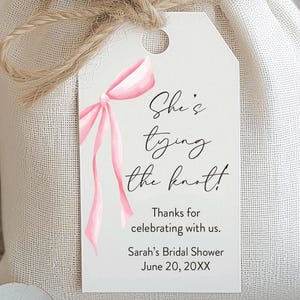 May include: A white gift bag tied with twine, featuring a tag with a pink watercolor bow and the text "She's tying the knot!" The tag also includes "Thanks for celebrating with us." and "Sarah's Bridal Shower June 20, 20XX."