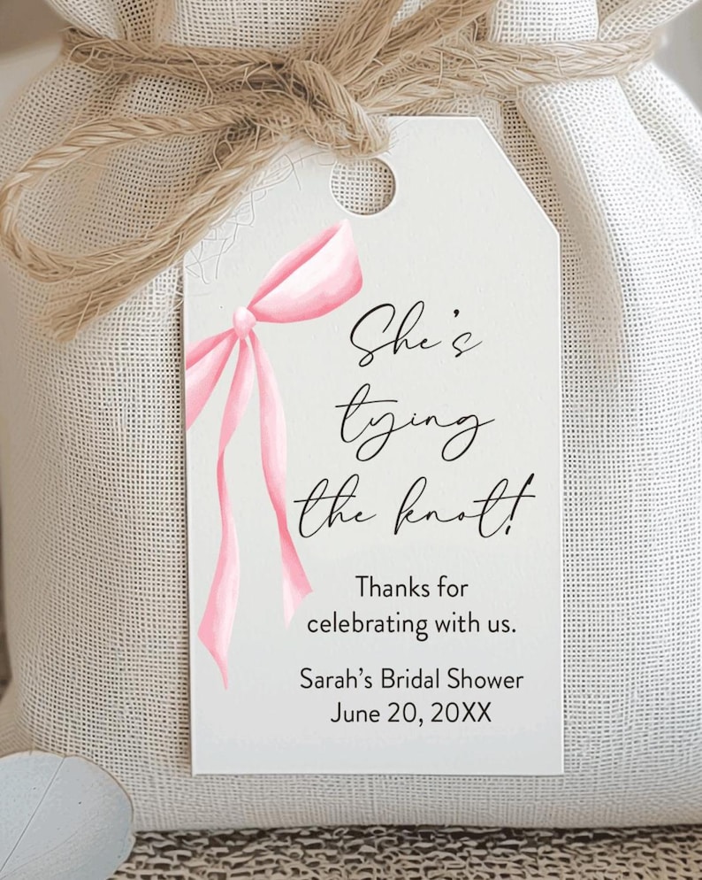 May include: A white gift bag tied with twine, featuring a tag with a pink watercolor bow and the text "She's tying the knot!" The tag also includes "Thanks for celebrating with us." and "Sarah's Bridal Shower June 20, 20XX."