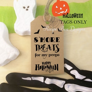 May include: A brown tag with the text "S'MORE TREATS for my peeps" and "Happy Halloween" with a spiderweb design. The tag is tied with twine and has a pumpkin and bats graphic. White ghost-shaped marshmallows and a skeleton hand are also in the image.