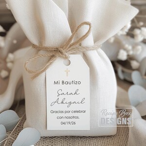 May include: A small, off-white fabric gift bag tied with twine. A white tag is attached with the text "Mi Bautizo Sarah Abigail Gracias por celebrar con nosotros. 04/19/26" and a cross symbol. The bag is from Recipe Box Designs.