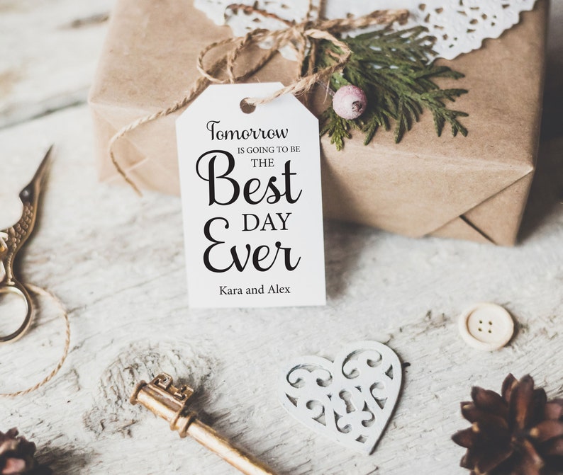 K&ouml;nnte beinhalten: Ein wei&szlig;es Geschenkanh&auml;nger mit schwarzem Text, der "Morgen wird der beste Tag aller Zeiten" lautet, mit den Namen Kara und Alex unten. Der Anh&auml;nger ist mit einer Schnur an einer braunen Geschenkbox gebunden.