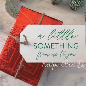 K&ouml;nnte beinhalten: Eine rechteckige Geschenkbox, eingewickelt in gl&auml;nzendes rotes Metallic-Papier mit einer Juteschnur. Daran befestigt ist ein wei&szlig;es rechteckiges Etikett mit gr&uuml;nem Text: a little SOMETHING from me to you. Der Text Recipe Box Designs ist rosa.