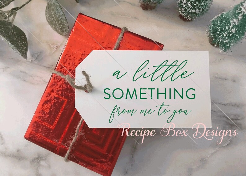K&ouml;nnte beinhalten: Eine rechteckige Geschenkbox, eingewickelt in gl&auml;nzendes rotes Metallic-Papier mit einer Juteschnur. Daran befestigt ist ein wei&szlig;es rechteckiges Etikett mit gr&uuml;nem Text: a little SOMETHING from me to you. Der Text Recipe Box Designs ist rosa.