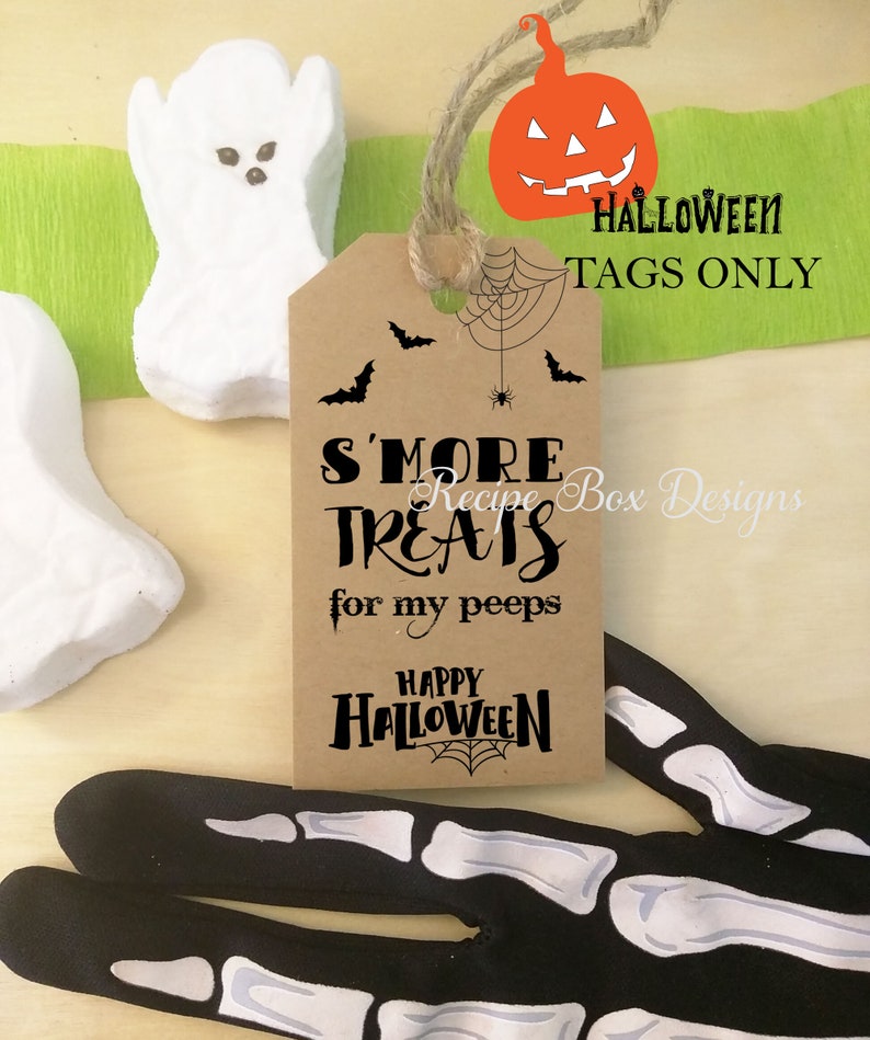 May include: A brown paper tag with the text "S'MORE TREATS for my peeps" and "Happy Halloween" with a spiderweb design. The tag is decorated with bats and a pumpkin. White ghost-shaped marshmallows and black skeleton gloves are also visible.