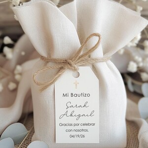 May include: A small, off-white fabric gift bag tied with twine. A white tag with a gold cross and the words "Mi Bautizo Sarah Abigail" is attached. The tag also includes the text "Gracias por celebrar con nosotros. 04/19/26".