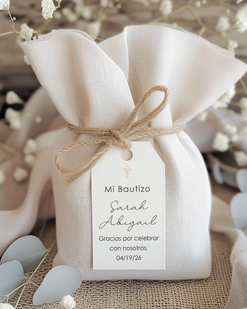 May include: A small, off-white fabric gift bag tied with twine. A white tag with a gold cross and the words "Mi Bautizo Sarah Abigail" is attached. The tag also includes the text "Gracias por celebrar con nosotros. 04/19/26".