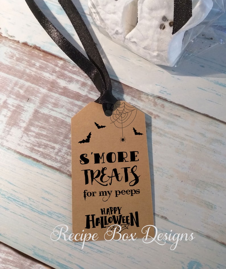 May include: A brown paper tag with black text and illustrations for Halloween. The tag reads "S'MORE TREATS for my peeps HAPPY HALLOWEEN" with bat and spider web graphics. A black ribbon is attached to the tag.