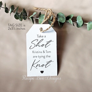 May include: White wedding favor tag with black text that reads "Take a Shot Kristina & Tom are tying the Knot". The tag is 2 by 3 1/2 inches and has a small black heart at the bottom. The tag is tied with twine and is sitting on a green branch.