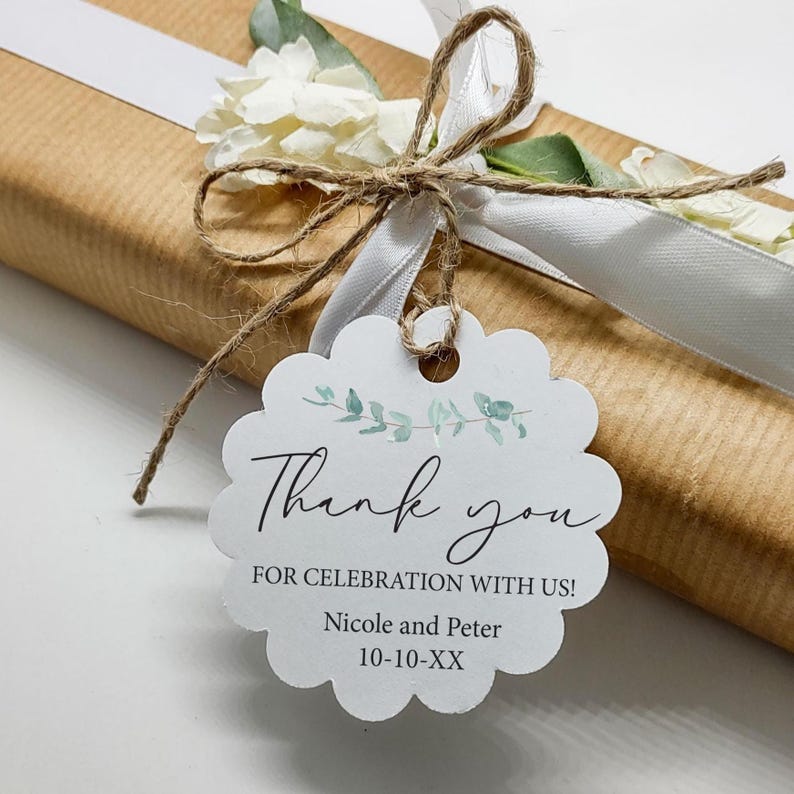 May include: A gift wrapped in brown paper, tied with a white ribbon and twine, and adorned with a white floral arrangement. A scalloped tag reads "Thank you" with a botanical design and names.