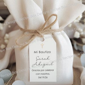 May include: A beige fabric gift bag tied with twine, featuring a white tag. The tag reads "Mi Bautizo" with the names "Sarah Abigail" and the date "04/19/26". A small cross is at the top of the tag.