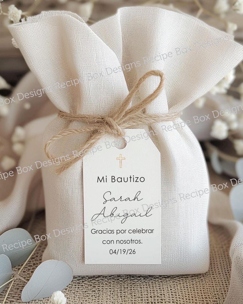 May include: A beige fabric gift bag tied with twine, featuring a white tag. The tag reads "Mi Bautizo" with the names "Sarah Abigail" and the date "04/19/26". A small cross is at the top of the tag.