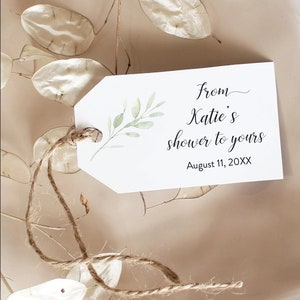 May include: A white gift tag with a green leafy branch design. The tag has the text "from Katie's shower to yours" and "August 11, 20XX". The tag is tied with a brown twine string.