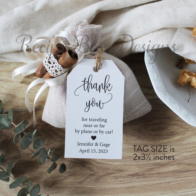 May include: A white thank you tag with black text. The tag says "thank you" in cursive, followed by "for traveling near or far by plane or by car!"  The tag also includes the names "Jennifer & Gage" and the date "April 15, 2023". The tag is tied to a small burlap bag filled with cinnamon sticks. The tag size is 2x3 1/2 inches.