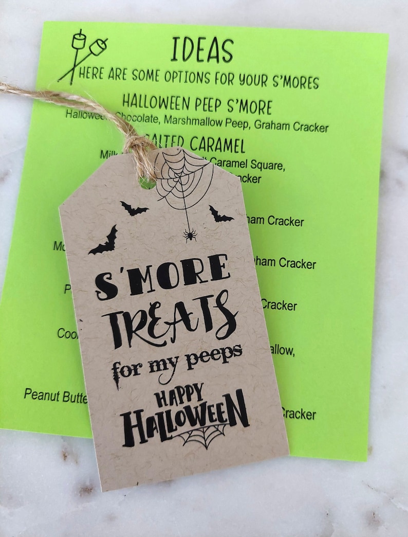 May include: A kraft paper tag with the text "S'MORE TREATS for my peeps HAPPY HALLOWEEN" is tied with twine to a neon green card with s'mores ideas. The tag features bats and a spider web design.