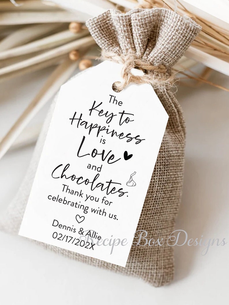 Puede incluir: Una etiqueta blanca con texto negro que dice "The Key to Happiness is Love and Chocolates." La etiqueta est&aacute; atada a una peque&ntilde;a bolsa de arpillera. El texto tambi&eacute;n incluye "Thank you for celebrating with us." y "Dennis & Allie 02/17/202X".