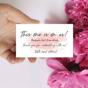 May include: A white card with the text "This one is on us!" in black script, with a pink watercolor-style background. The card also says "Redeem for 1 free drink" and "Thank you for celebrating with us!" with the names "Kate and Grant".