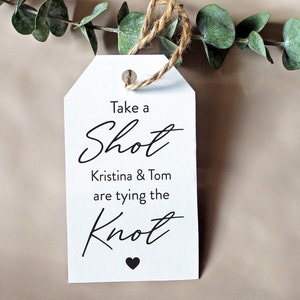 May include: White tag with a brown twine string attached. The tag has black text that reads "Take a Shot Kristina & Tom are tying the Knot" with a small black heart at the bottom.