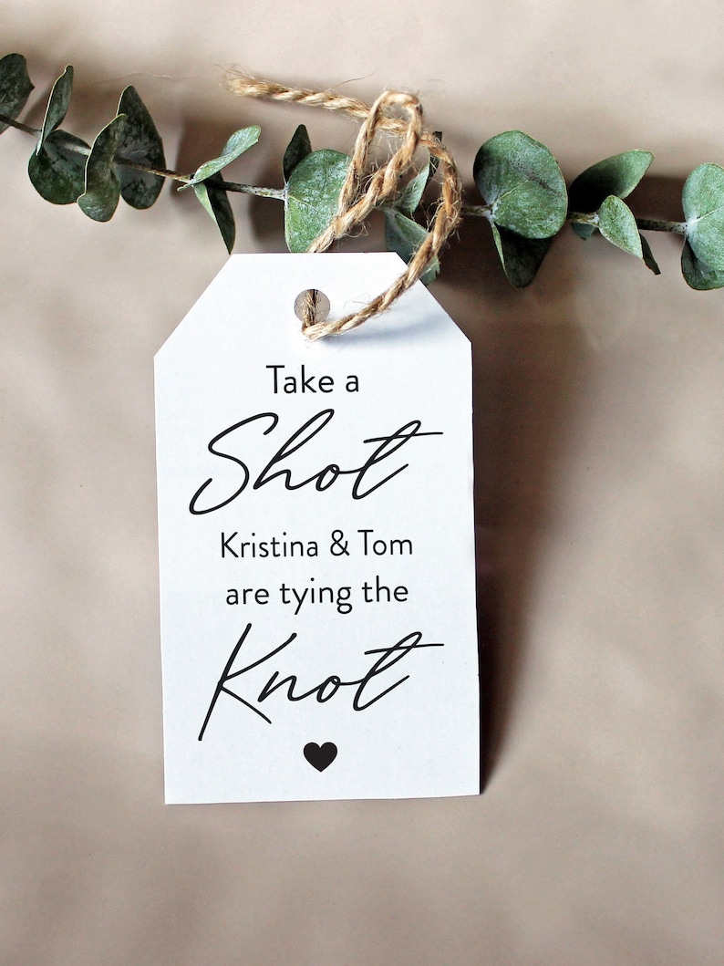 May include: White tag with a brown twine string attached. The tag has black text that reads "Take a Shot Kristina & Tom are tying the Knot" with a small black heart at the bottom.