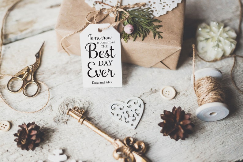 K&ouml;nnte beinhalten: Ein wei&szlig;es Geschenkanh&auml;nger mit schwarzem Text, der "Morgen wird der beste Tag aller Zeiten, Kara und Alex" lautet. Der Anh&auml;nger ist an einer mit braunem Papier verpackten Geschenkbox mit Schnur und einem Zweig Gr&uuml;n gebunden. Die Geschenkbox ist umgeben von anderen Bastelutensilien, darunter eine Schnurspule, eine herzf&ouml;rmige Ausschneidung und Tannenzapfen.