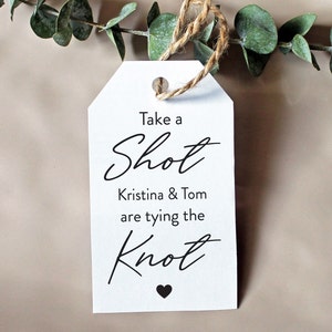May include: White tag with black text that reads "Take a Shot Kristina & Tom are tying the Knot" with a small black heart at the bottom.