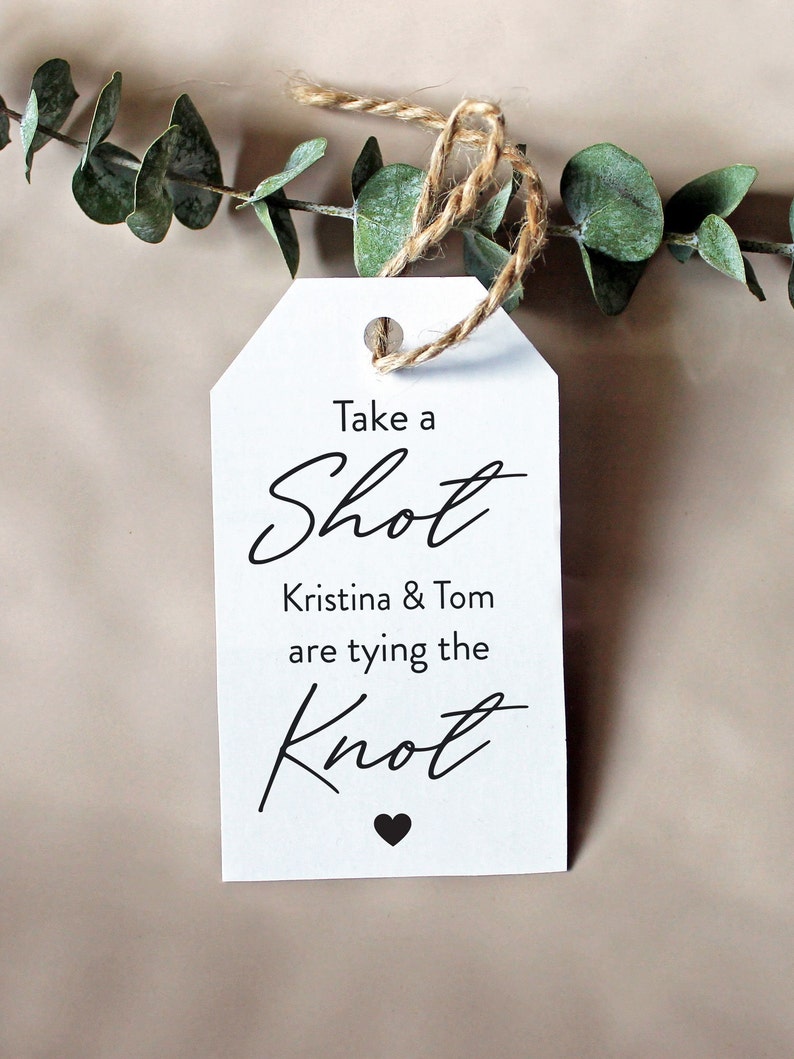 May include: White tag with black text that reads "Take a Shot Kristina & Tom are tying the Knot" with a small black heart at the bottom.