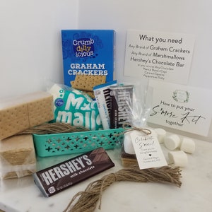 May include: A teal basket filled with ingredients for making s'mores. The basket contains graham crackers, marshmallows, a Hershey's chocolate bar, and a small tag that says "Celebrate S'more!" The basket is sitting on a white surface with a white card that says "What you need" and a white card that says "How to put your S'more Kit together".