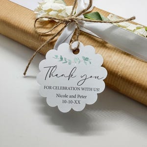 May include: A wrapped gift in brown paper, tied with a white ribbon and twine. A white scalloped tag with the words "Thank you" and "For celebration with us!" is attached. The tag also includes names and a date.