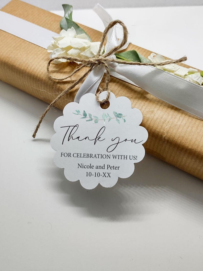 May include: A wrapped gift in brown paper, tied with a white ribbon and twine. A white scalloped tag with the words "Thank you" and "For celebration with us!" is attached. The tag also includes names and a date.