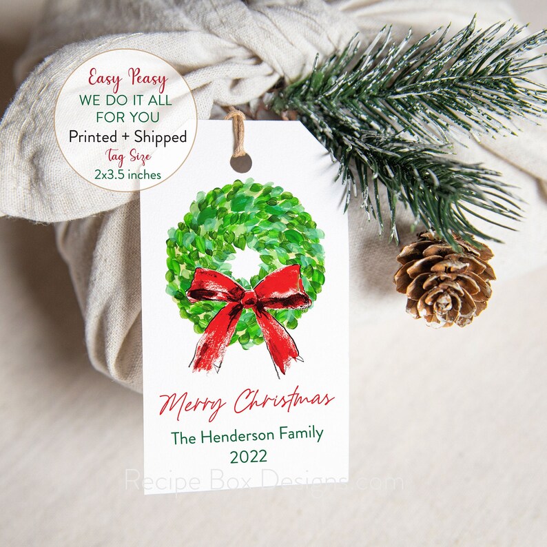 May include: A white gift tag with a watercolor wreath and red bow. The tag reads "Merry Christmas" and "The Henderson Family 2022." The tag is 2 x 3.5 inches. A sprig of evergreen and a pinecone are next to the tag.