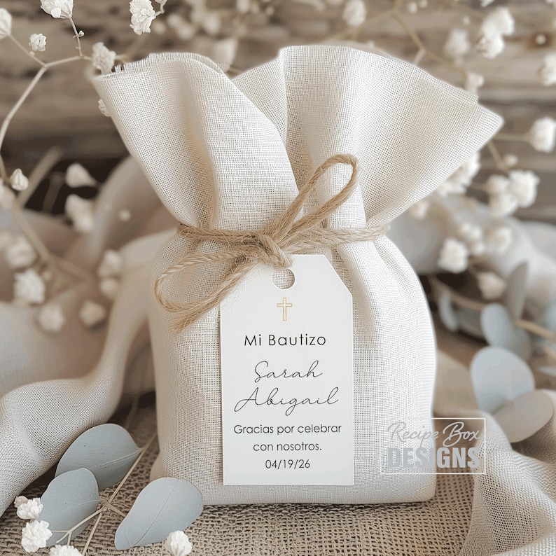 May include: A small, off-white fabric gift bag tied with twine, featuring a white tag with the text "Mi Bautizo Sarah Abigail" and a cross symbol. The tag also includes the date 04/19/26 and the phrase "Gracias por celebrar con nosotros."