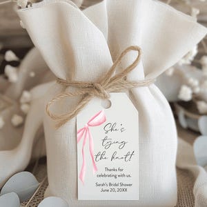 May include: A small, beige fabric gift bag tied with twine, featuring a white tag with pink ribbon art and the text "She's tying the knot!" and "Sarah's Bridal Shower June 20, 20XX."