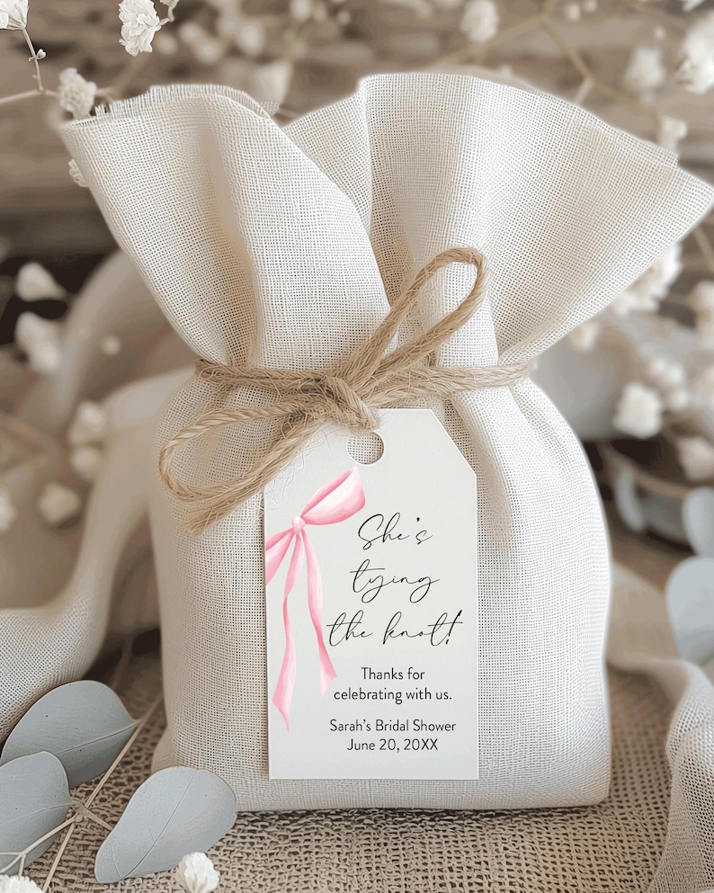 May include: A small, beige fabric gift bag tied with twine, featuring a white tag with pink ribbon art and the text "She's tying the knot!" and "Sarah's Bridal Shower June 20, 20XX."