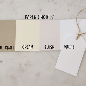 Może przedstawiać: Cztery pr&oacute;bki papieru oznaczone jako "Light Kraft", "Cream", "Blush" i "White". Pr&oacute;bki są ułożone od lewej do prawej, a pr&oacute;bka "White" to etykieta wisząca na sznurku.
