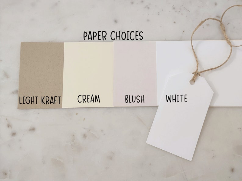 Może przedstawiać: Cztery pr&oacute;bki papieru oznaczone jako "Light Kraft", "Cream", "Blush" i "White". Pr&oacute;bki są ułożone od lewej do prawej, a pr&oacute;bka "White" to etykieta wisząca na sznurku.