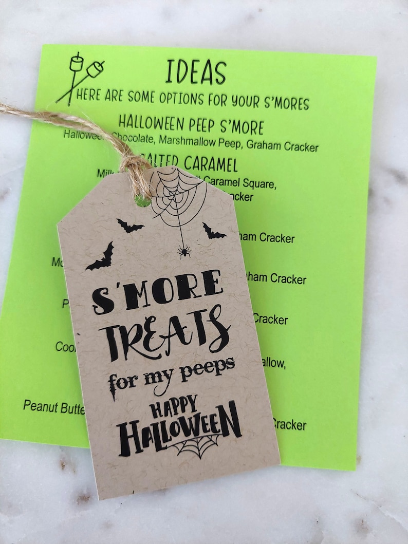 May include: A kraft paper tag with the text "S'MORE TREATS for my peeps HAPPY HALLOWEEN" and a spiderweb design, hanging over a bright green card with "IDEAS" and s'mores recipe suggestions. A twine string is attached.