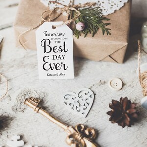 K&ouml;nnte beinhalten: Ein wei&szlig;es Geschenkanh&auml;nger mit schwarzem Text, der "Morgen wird der beste Tag aller Zeiten. Kara und Alex." lautet. Der Anh&auml;nger ist mit einer Schnur an einer braunen Geschenkbox gebunden. Die Geschenkbox ist mit einer wei&szlig;en Spitzendeckchen und einem gr&uuml;nen Zweig verziert.