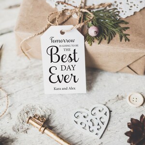K&ouml;nnte beinhalten: Ein wei&szlig;es Geschenkanh&auml;nger mit schwarzem Text, der "Morgen wird der beste Tag aller Zeiten" lautet, mit den Namen Kara und Alex unten. Der Anh&auml;nger ist mit einer Schnur an einer braunen Geschenkbox gebunden.