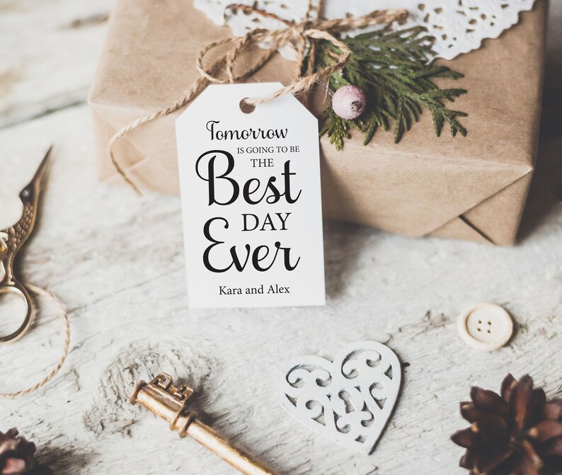 K&ouml;nnte beinhalten: Ein wei&szlig;es Geschenkanh&auml;nger mit schwarzem Text, der "Morgen wird der beste Tag aller Zeiten" lautet, mit den Namen Kara und Alex unten. Der Anh&auml;nger ist mit einer Schnur an einer braunen Geschenkbox gebunden.