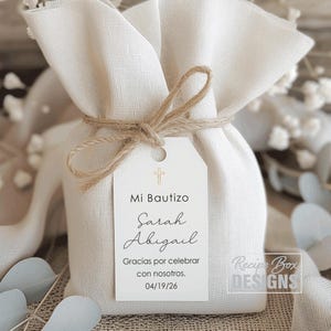 May include: A small, off-white fabric gift bag tied with twine. A white tag is attached with the text "Mi Bautizo" and the names "Sarah Abigail". The tag also includes the date 04/19/26 and the phrase "Gracias por celebrar con nosotros."