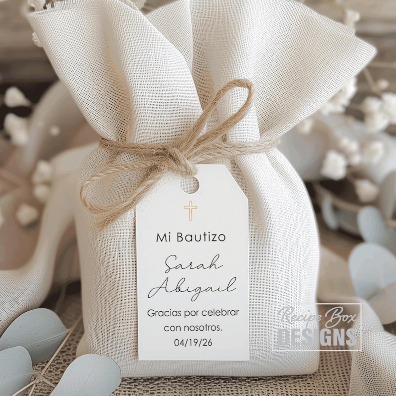May include: A small, off-white fabric gift bag tied with twine. A white tag is attached with the text "Mi Bautizo" and the names "Sarah Abigail". The tag also includes the date 04/19/26 and the phrase "Gracias por celebrar con nosotros."